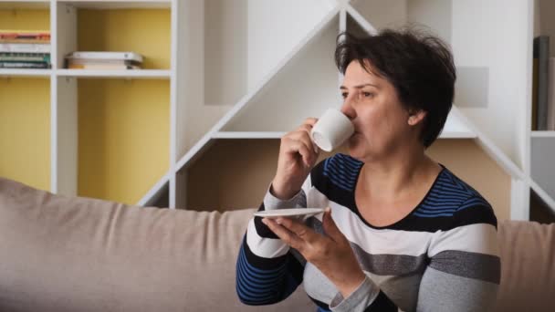 Kobieta pije filiżankę kawy, siedzi na kanapie w domu — Wideo stockowe