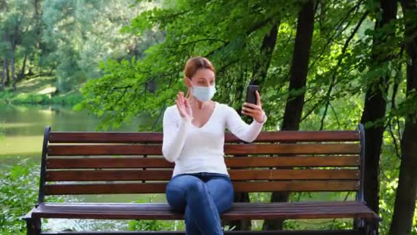 Junge Frau sitzt mit Maske allein in einem Park und spricht während der Epidemie über einen Videokanal. Sicherheit, Entfernung, Gesundheit, N1H1, Virenschutz, Covid, Viren- und Coronavirus-Konzept — Stockvideo