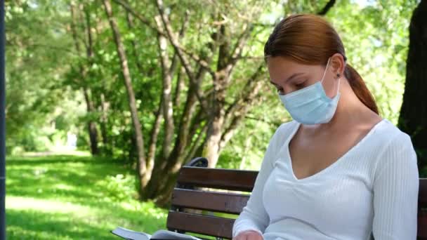 Junge Frau sitzt mit Maske allein in einem Park und liest während der Epidemie ein Buch. Sicherheit, Entfernung, Gesundheit, N1H1, Virenschutz, Covid, Viren- und Coronavirus-Konzept — Stockvideo