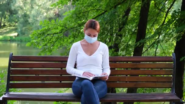 Νεαρή γυναίκα κάθεται μόνη σε ένα πάρκο με μάσκα και διαβάζει ένα βιβλίο κατά τη διάρκεια της επιδημίας. Ασφάλεια, απόσταση, υγεία, N1H1, προστασία από ιούς, covid, ιός και coronavirus έννοια — Αρχείο Βίντεο