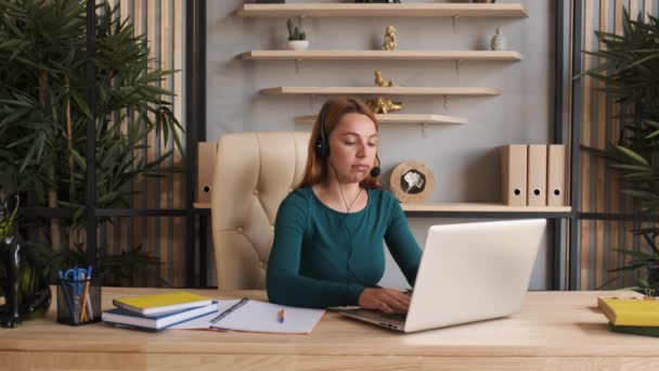 若い女性は、会議の呼び出しによって通信ヘッドセットを着用自宅のオフィスでコンピュータを見て話す、ビデオチャットジョブインタビューやオンライン教師の概念との距離言語コースクラス — ストック動画