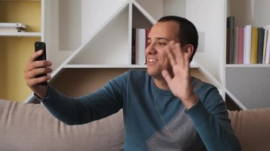 Akıllı telefonlu bir adam internetten cep telefonuyla arıyor evdeki kanepede ekrana bakıyor ve selam veriyor.