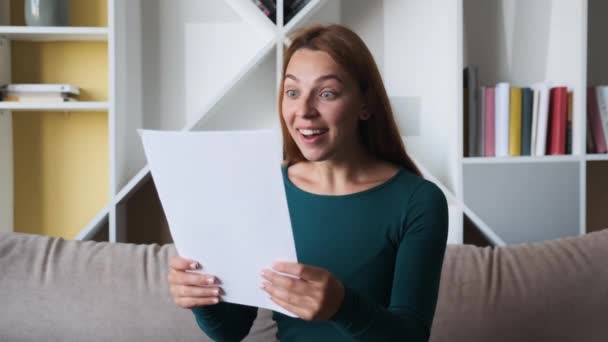 幸せな若いアメリカ人女性は、良いニュースに驚いて、自宅でソファの上に座って紙の手紙を読む。良い銀行ニュースを受け取ることを祝う喜びの出産の女性、最後の住宅ローンの支払い. — ストック動画