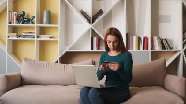 Pretty girl dokonuje płatności online trzymając kartę bankową za pomocą nowoczesnego laptopa w domu siedzi na kanapie. Finanse, zakupy w internecie i koncepcja młodzieży. — Wideo stockowe