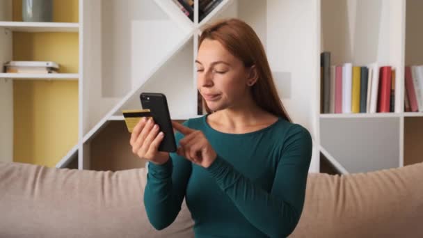 Kredi kartı ve akıllı telefon tutan genç bayan müşteri evdeki kanepede oturuyor. İnternetten alışveriş yapan mutlu bayan alışverişçi kolay taşınabilir ödemeler yapıyor. E-bankacılık uygulama servisi — Stok video