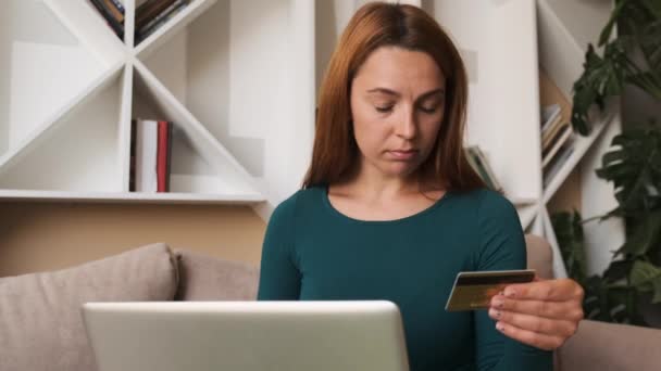 Красивая девушка делает онлайн-платеж держа банковскую карту, используя современный ноутбук дома, сидя на диване. Финансы, покупки в Интернете и молодежной концепции. — стоковое видео