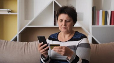 Kredi kartı ve akıllı telefon tutan yaşlı bir kadın evdeki koltukta oturuyor. İnternetten alışveriş yapan mutlu bayan alışverişçi kolay taşınabilir ödemeler yapıyor. E-bankacılık uygulama servisi