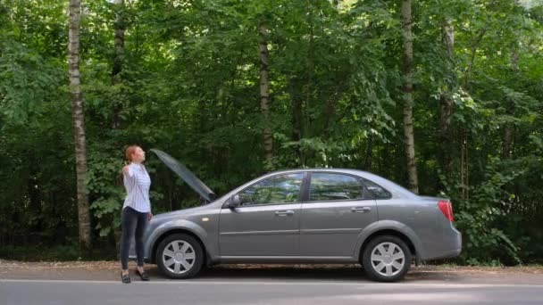 Женщина пытается остановить машину на дороге. Напряженная женщина-водитель стоит перед открытым капотом. Ошибка, проблема. — стоковое видео