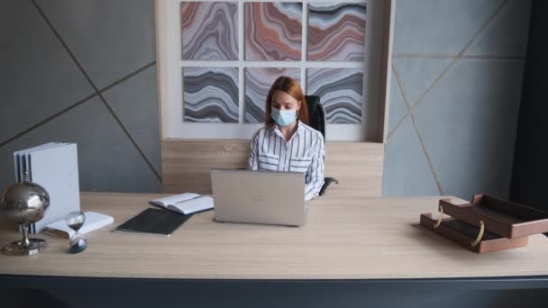 Trabajo durante el auto-aislamiento y cuarentena. Una joven empleada con una máscara facial médica trabaja en la oficina del ordenador. Covid Pandémico del Coronavirus 19. — Vídeos de Stock