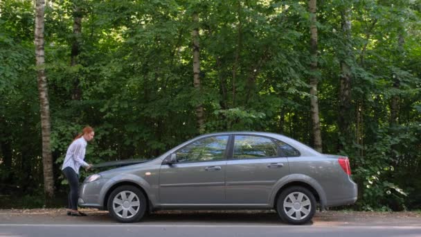 Vue latérale de femme confiante ouvrant le capot de la voiture et regardant le moteur. Une conductrice debout à côté d'un véhicule cassé près de la forêt. Accident de voiture sur route rurale. — Video