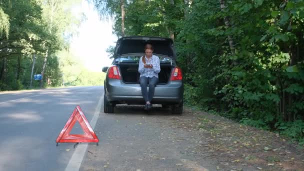 女性は車のトランクに座って、森の中で壊れた車を助けるために携帯電話を使用しています。非常停止標識として赤の三角形。道路上の車の事故. — ストック動画