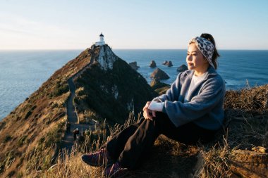 Nugget Point, Dunedin, Yeni Zelanda 'da takılan Asyalı güzel bir turist. Genç Asyalı gezgin sabah Yeni Zelanda kıyılarında kahvenin tadını çıkarıyor. Doğal ortamdaki insanların yaşam tarzı..