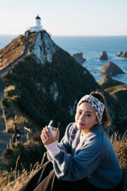 Beautiful asian tourist chilling out at Nugget Point, Dunedin, New Zealand. Young asian traveller enjoys coffee in morning at coastline of New Zealand. Lifestyle image of people in natural landscape. clipart