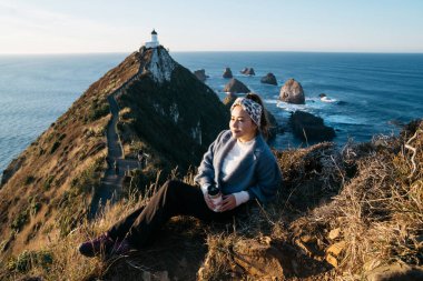 Nugget Point, Dunedin, Yeni Zelanda 'da takılan Asyalı güzel bir turist. Genç Asyalı gezgin sabah Yeni Zelanda kıyılarında kahvenin tadını çıkarıyor. Doğal ortamdaki insanların yaşam tarzı..