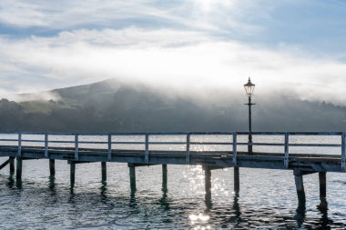 Yeni Zelanda 'daki Akaroa limanında ahşap iskele ve yelkenli. Birkaç beyaz bulut ve sis ile açık bir gökyüzü. Rahatlamanın, zenginliğin, boş zamanların sembolü. Panorama biçimi.
