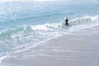 Sörf tahtasıyla dalış kıyafeti giyen sörfçü, Yeni Zelanda, Dunedin 'deki St. Clair plajındaki kayaların üzerindeki okyanus dalgalarını seyrediyor..