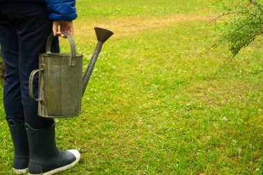 Elinde metal bir su bidonu olan lastik çizmeli bir çocuk bahçedeki yeşil çimlerin arka planına yaslanmış..