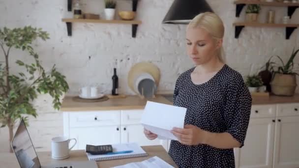 Молода жінка, стоячи на кухні, відкриває рахунок за комунальні послуги і дуже засмучена — стокове відео