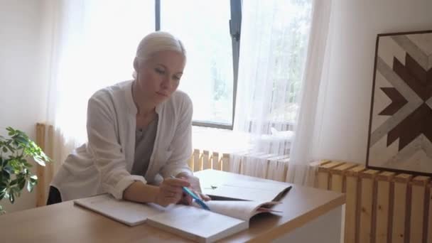 Studentin studiert zu Hause am Tisch. Macht Notizen aus einem Buch in einem Notizbuch — Stockvideo