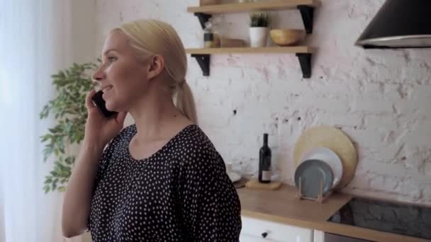 Una guapa rubia está hablando por teléfono, parada en la cocina luminosa. — Vídeos de Stock