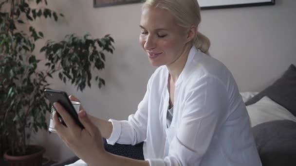 Jovem dona de casa na cama conversando com um amigo em um vídeo usando seu smartphone — Vídeo de Stock