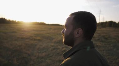 Sakallı genç bir adam sonbahar tarlasında gün batımında yürüyor ve hayatın tadını çıkarıyor.