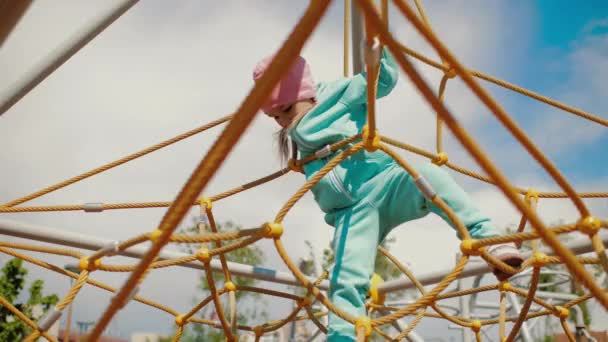 Menina bonito rasteja na estrutura de rede de escalada no parque infantil segurando em cordas — Vídeo de Stock