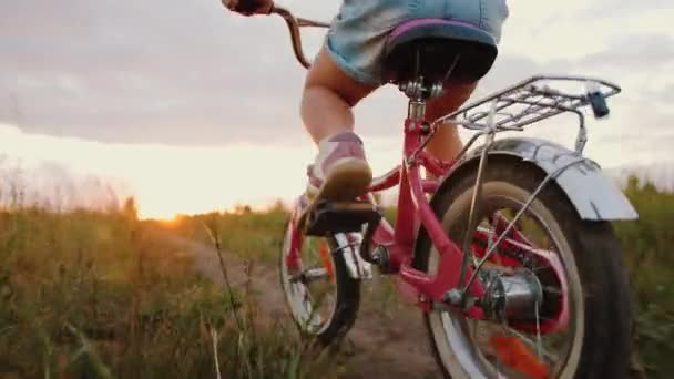Kislány lovaglás kerékpár a réten nyomvonal naplementekor, pedál közelről — Stock videók