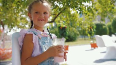 5-6 yaşlarında bir kız dışarıda bir kafede oturmuş milkshake içiyor ve kameraya gülümsüyor.