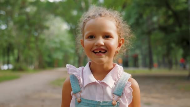 Portret van klein schattig meisje kijkt naar camera, glimlacht en lacht in de zomer park — Stockvideo