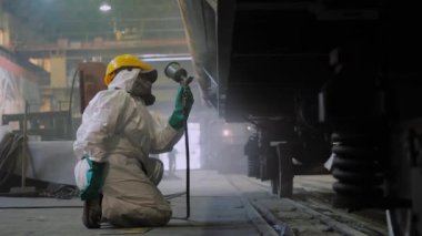 Koruyucu giysili fabrika işçisi tren vagonunun yüzeyini sprey silahla boyuyor.
