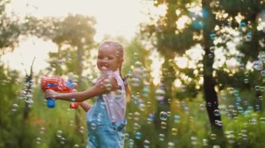 Küçük mutlu kız sabun köpüğü üflüyor, parkta oyuncak tabancayla oynuyor.