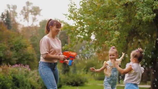 Mãe feliz soprando bolhas de sabão com arma de brinquedo, filhas pequenas tentando pegar — Vídeo de Stock