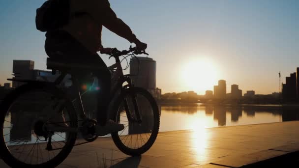 Siluetas de dos ciclistas cabalgando en el terraplén del estanque de la ciudad al atardecer — Vídeos de Stock
