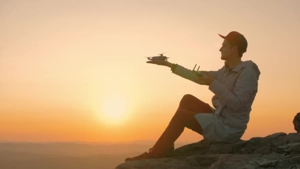 Hombre sentado en el borde del acantilado de roca y comienza a dron de su mano al atardecer — Vídeo de stock
