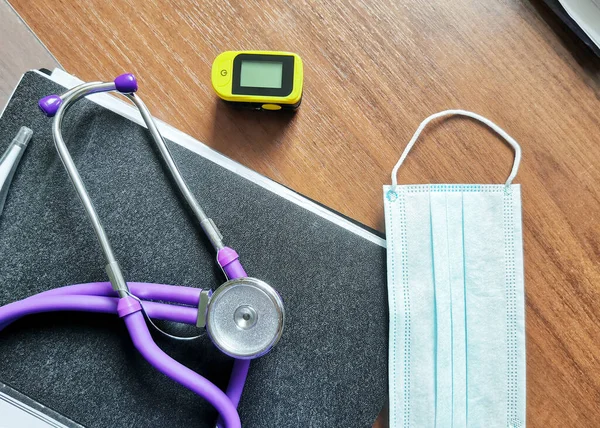 一个黑色塑料文件夹上的听诊器 一个指尖脉动血氧计和医疗面罩躺在一张木制桌子上 顶部视图 — 图库照片