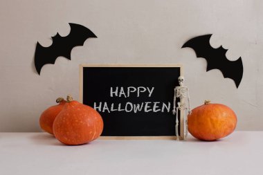 Tebeşir tahtası, iskelet, yarasalar, beyaz masada turuncu balkabakları, beton arka plan görünüm kopyalama alanı. Cadılar Bayramı kutlaması. Ürkütücü dekorasyonlar. Gece partisi. Ölüler Günü