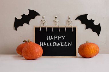 Tebeşir tahtası, iskelet, yarasalar, beyaz masada turuncu balkabakları, beton arka plan görünüm kopyalama alanı. Cadılar Bayramı kutlaması. Ürkütücü dekorasyonlar. Gece partisi. Ölüler Günü