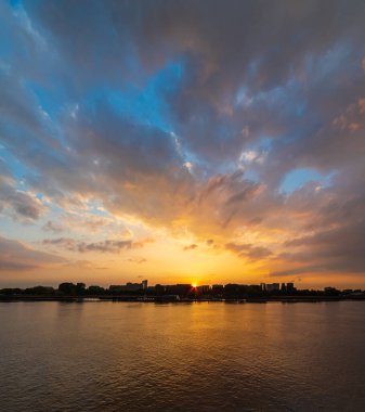 Gün batımında bir nehirde şehrin silueti. Görkemli altın gökyüzünün altında. Binaların arkasında güneş batıyor ve bir sürü kopya alanı var.
