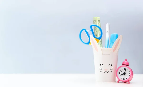 School supplies in paper cup in form of rabbit, pink clock on white and gray backdrop. Back to school. Stationery. Space for text.