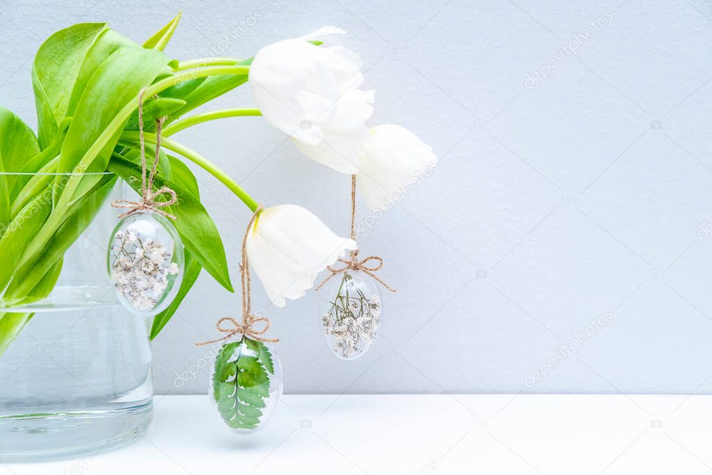 Easter minimal composition, white tulips, transparent glass eggs filled with white flowers and fern leaves on light grey background. Eco Stylish decor concept. Copy space. Festive greeting card, front view