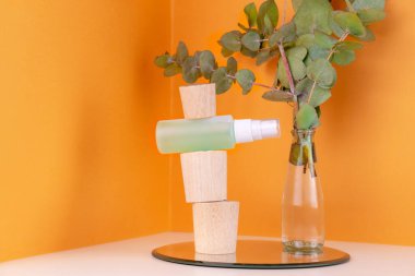 Ahşap kaidede yeşil sprey tonik şişesi ve turuncu arka planda cam vazoda okaliptüs dalı. Kozmetik ürünleri. Deri iyileşmesi için organik doğal tedavi