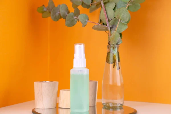 Green spray skin tonic bottle on wooden pedestal and eucalyptus branch in glass vase on orange backdrop. Care cosmetics. Organic natural treatment for skin healing