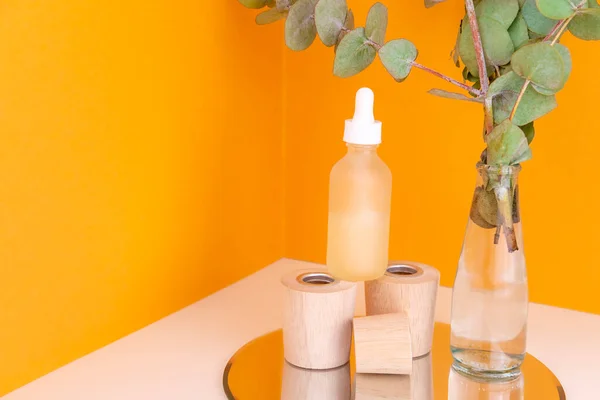Ahşap kaidede Retinol serumu şişesi ve turuncu arka planda okaliptüs dalı olan cam vazo. Organik iyileştirme kozmetikleri. Eko cilt bakımı. Güzel hyaluronik asit tedavisi