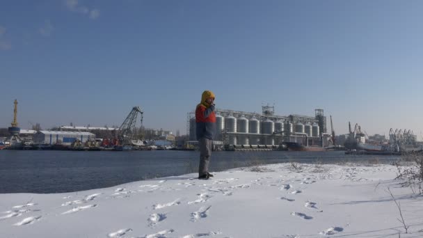 男性労働者はポートの穀倉の産業ビューの背景を冬の川の銀行に沿って歩いてください — ストック動画