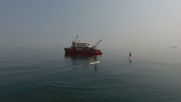 空中风景 两名男子运动员带着桨登上冲浪板 靠近双桅帆船 远离大海 在平静的水面上航行 — 图库视频影像