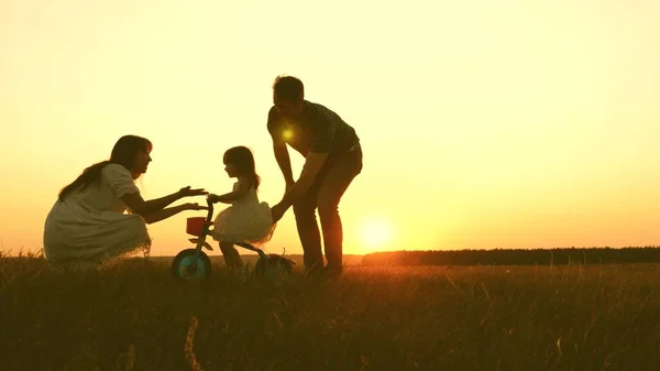 Mom and dad teach a young child to ride a bike at sunset in the park. Teamwork. Silhouette family walk — Stock Photo, Image