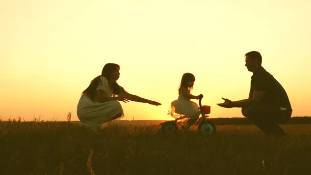 Maman et papa apprennent à un jeune enfant à faire du vélo au coucher du soleil dans le parc. Travail d'équipe. Silhouette promenade familiale — Video