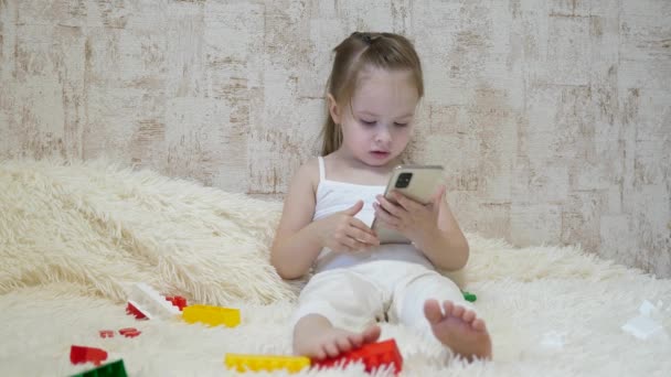 Ein Kind spielt am Telefon, während es mit dem Designer auf der Couch sitzt. Spielzeug im Smartphone. Das Leben als Fernvorschulerziehung. Modernes Baby — Stockvideo