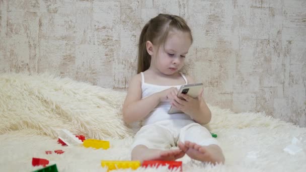 Kind spielt am Telefon, während es mit dem Designer auf der Couch sitzt. Spielzeug im Smartphone. Das Leben als Fernvorschulerziehung. Modernes Baby — Stockvideo